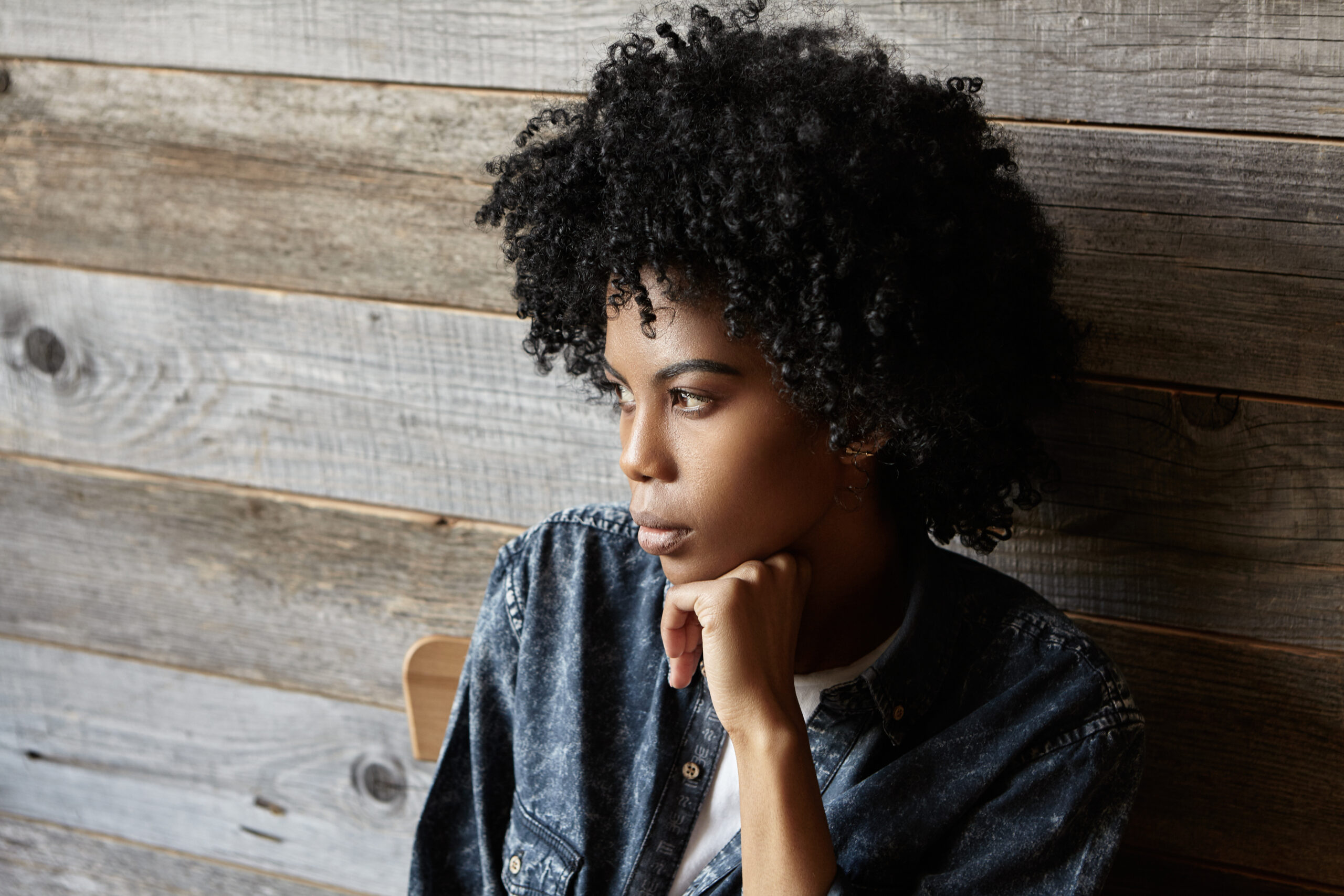 Portrait Of Fashionable Young African American Female Dressed In Denim Shirt Having Serious And Thoughtful Look, Thinking About Something Important, Sitting At Cafe With Head Reclined Upon Her Hand